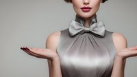 Elegant young Caucasian woman in silver sleeveless top with bow tie collar presenting with open palms, professional business portrait on neutral background.