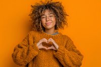 Young woman with curly hair and glasses making heart shape with hands against vibrant orange background, wearing matching knit sweater.
