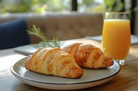 Fresh croissants with rosemary garnish and orange juice on plate, morning breakfast setup on wooden table near window for cafe menu design.