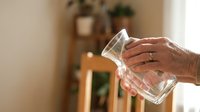 Senior woman hands holding empty clear glass cup in cozy home interior with natural lighting and plants in background for hydration concept.