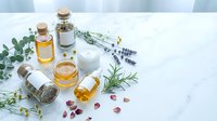 Natural spa aromatherapy setup with essential oil bottles, cream jar, herbs and flowers on white marble surface for wellness and relaxation treatments.