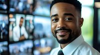 Professional African American businessman smiling confidently in modern office environment with video conference screens displaying remote team members in background.