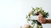 Hands holding blank white card with fresh flower arrangement including roses and eucalyptus greenery on white background for greeting, invitation, or promotional messaging.