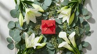 Elegant white lilies and eucalyptus branches surrounding red gift box on white fabric background for holiday celebrations and special occasions.
