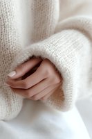 Female hand with neutral manicure emerging from soft white knitted sweater sleeve, creating cozy winter mood. Gentle fashion detail for lifestyle blogs and social media.