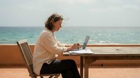 Middle-aged woman with curly brown hair, Caucasian ethnicity, working remotely on laptop at wooden table overlooking turquoise ocean. Freelancer or digital nomad concept for remote work, travel lifestyle, and flexible employment opportunities.