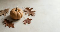 Small orange pumpkin with dried autumn oak leaves on neutral beige background for seasonal decoration and fall harvest themed designs.