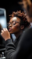 Young African American woman with natural curly hair and glasses applying makeup while looking in mirror, wearing casual black attire. Professional beauty routine moment.