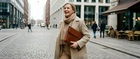 Professional blonde businesswoman walking confidently through urban street carrying leather portfolio, wearing elegant beige coat for corporate lifestyle.