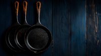 Cast iron skillets hanging on dark blue wooden wall with dramatic lighting and rustic atmosphere, minimalist kitchen decor for food photography and restaurant design.