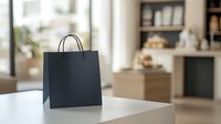 Black paper shopping bag on white table in modern interior with blurred background. Minimalist retail and consumer concept for design layouts.