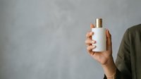 Male hand holding white cosmetic bottle with gold cap against gray background for skincare product marketing.