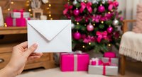 Hand holding white envelope with Christmas tree and pink gift boxes in cozy holiday living room background for seasonal greeting cards and invitations.
