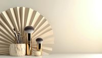 Makeup brushes with golden handles against geometric paper fan backdrop in neutral beige tones, minimal cosmetic still life composition on white surface.