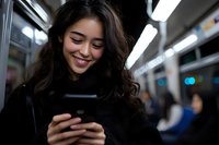 Young Asian woman with long dark hair smiling while using smartphone on subway train. Blurred urban transport interior and lights in background.