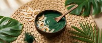 Green spirulina smoothie bowl with granola toppings on wicker placemat surrounded by tropical monstera leaves for healthy breakfast concept.