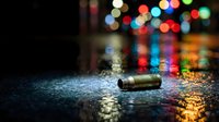 Empty bullet shell casing on wet asphalt street at night with colorful bokeh lights reflecting on pavement creating dramatic urban crime scene atmosphere.