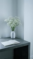 Minimalist workspace with white gypsophila flowers in a ceramic vase and open notebook on a concrete desk, soft neutral tones.