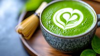 Green matcha latte with white heart-shaped foam art in traditional ceramic cup on wooden surface, bamboo whisk and fresh leaves visible.