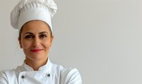 Professional female chef in white uniform and traditional toque smiling warmly against light background, expressing confidence and expertise in culinary arts.