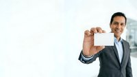 Middle-aged man with South Asian ethnicity wearing dark business suit and light blue shirt, smiling while holding blank white business card toward camera in professional office setting.