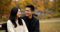 Happy Asian couple sitting on park bench during autumn season sharing intimate moment together outdoors with colorful fall foliage background for lifestyle content.