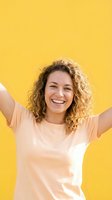 Young woman with curly golden-brown hair and fair complexion wearing a peach t-shirt, arms raised in celebration against a bright yellow background, expressing joy and positivity for lifestyle and wellness campaigns.