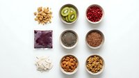 Healthy superfood ingredients in bowls on white background including nuts, seeds, berries and greens for nutritious meal preparation.