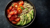 Fresh quinoa bowl with sliced ripe avocado, juicy tomatoes and black sesame seeds served in dark ceramic bowl on black stone background.