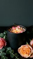 Luxury scented candle with dried flowers and rose petals in black ceramic holder surrounded by fresh pink roses for spa relaxation and aromatherapy.