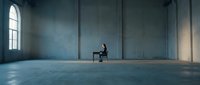 Solitary figure seated on chair in vast empty industrial loft space with concrete walls and natural light from tall windows, conveying isolation and contemplation.