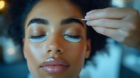 Young woman with medium skin tone receiving professional eyeshadow application, blue cosmetic product applied to eyelids during makeup tutorial or beauty treatment session.