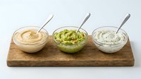 Assorted dips and spreads in bowls with spoons on wooden serving board, including hummus, guacamole, and tzatziki for appetizers and entertaining.