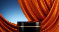 Black cylindrical podium with flowing orange silk fabric drapes against blue gradient background for luxury product display and presentation.