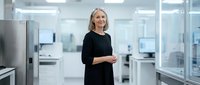 Mature woman with blonde hair wearing black clothing standing in modern laboratory or medical facility with glass partitions and equipment.