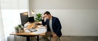 Stressed businessman holding head in hands at modern office desk with laptop and documents showing work pressure and mental fatigue.