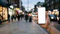 Male hand holding smartphone with blank white screen in urban shopping street at dusk, displaying mobile app interface for retail, commerce, or technology marketing purposes.