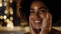 Young African American woman with natural curly hair smiling warmly while touching her face, glowing skin illuminated by soft bokeh lights creating magical evening atmosphere.