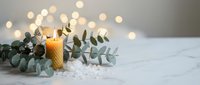 Natural beeswax candle with eucalyptus branches and sea salt on white marble surface with warm bokeh lights creating cozy spa atmosphere for relaxation.