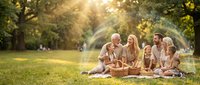 Multi-generational family enjoying picnic in sunny park with golden sunlight filtering through trees, creating warm atmosphere for outdoor gathering.