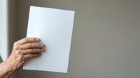 Senior adult hand holding blank white paper sheet against neutral wall background for mockup and design purposes.