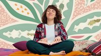 Young woman with brown wavy hair sitting cross-legged reading book against colorful botanical wall mural. Student in casual plaid shirt enjoying literature indoors.