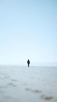 Solitary figure in dark wetsuit walking on empty sandy beach toward calm ocean waters under overcast sky, conveying solitude and contemplation.