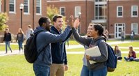 Diverse college students celebrating success with high five on university campus lawn during sunny day with brick buildings in background.