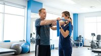 Personal trainer assisting young woman with resistance band exercise in modern fitness center for strength training and rehabilitation.