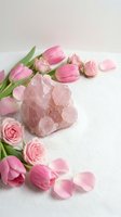 Rose quartz crystal cluster surrounded by pink tulips and roses on white surface, soft romantic aesthetic for wellness and beauty concepts.