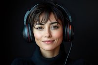 Young Caucasian woman wearing black sweater and gaming headphones smiles at camera against dark background, professional studio portrait with dramatic lighting.