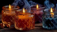 Aromatic scented candles in colorful glass jars with diamond pattern, burning with golden flames and wisps of smoke against dark background.