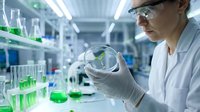 Female scientist in white lab coat and safety glasses examining green liquid sample in petri dish during laboratory research experiment.