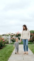Young mother walking with toddler son on garden path surrounded by blooming roses and white picket fence in suburban backyard setting.